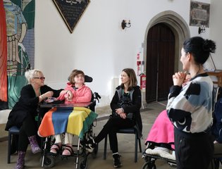 Woman in a wheelchair looking at a Yoga therapist who is standing with her hands held together.