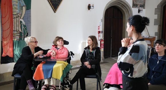 Woman in a wheelchair looking at a Yoga therapist who is standing with her hands held together.
