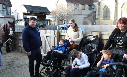 3 young boys in wheelchairs with 3 adults in a horse riding paddock