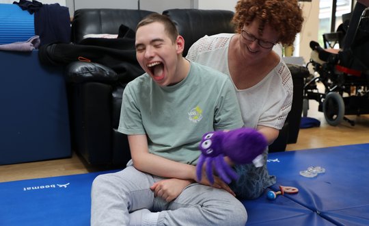 Boy smiling while sitting on the floor with a purple teddy bear.