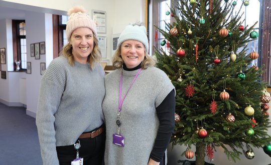 Two ladies smiling with woolly hats in front of a Christmas tree.